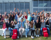 Vor dem modernen Gebäude mit den großen Fenstern der Hochschule Coburg hat sich eine Gruppe von Menschen im Freien versammelt. Sie lächeln und heben jubelnd die Hände, flankiert von zwei roten Blöcken mit weißen Buchstaben.