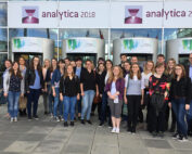 Eine Gruppe junger Erwachsener steht lächelnd vor dem Eingang zur Analytica 2018, einem Konferenzzentrum, über dessen Türen ein Schild prangt. Sie sind leger gekleidet und scheinen Studenten der Hochschule Coburg zu sein, die gespannt darauf sind, die neuesten Erkenntnisse der Branche zu erfahren.