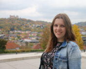 Eine Frau mit langen braunen Haaren lächelt in die Kamera. Sie trägt eine Jeansjacke über einem geblümten Oberteil. Sie steht vor einem malerischen Blick auf die Hochschule Coburg, mit einer Stadt am Hang und einem Schloss in der Ferne unter einem bewölkten Himmel.