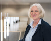 Eine ältere Frau mit kurzen weißen Haaren steht lächelnd in einem modernen, gut beleuchteten Innenraum der Hochschule Coburg. Sie trägt einen dunklen Blazer über einer hellblauen Bluse, was ihr einen professionellen Touch verleiht. Hängelampen und eine Treppe sorgen im Hintergrund für eine einladende Atmosphäre.