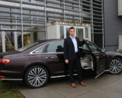 Ein Mann im Anzug steht neben der offenen Autotür einer Luxuslimousine, die auf einem gepflasterten Platz in der Nähe der Hochschule Coburg geparkt ist. Das Gebäude dahinter ist mit großen Fenstern und Metallpaneelen versehen. Das Auto ist dunkelbraun mit silbernen Felgen und strahlt vor der akademischen Kulisse Eleganz aus.