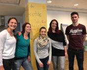 Eine Gruppe von fünf Personen, leger gekleidet, steht drinnen neben einer gelben Säule mit Schrift. Vier Frauen und ein Mann in einem kastanienbraunen T-Shirt lächeln herzlich. Die Umgebung erinnert an einen Unterrichts- oder Besprechungsraum der Hochschule Coburg.