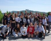 Eine Gruppe lächelnder Menschen, überwiegend Teenager, posiert gemeinsam im Freien vor einer Hügellandschaft mit einem Schloss im Hintergrund. In legerer Kleidung unter dem sonnigen Himmel verkörpern sie den lebendigen Geist, der oft an der Hochschule Coburg herrscht.