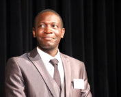 Ein Mann in Anzug und Krawatte steht vor einem dunklen Vorhang, blickt leicht zur Seite und hat einen neutralen Gesichtsausdruck. An seiner Jacke trägt er ein Namensschild, auf dem dezent „Hochschule Coburg“ steht.