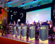 Bei einem Big-Band-Auftritt auf der Bühne spielen talentierte Musiker der Hochschule Coburg Saxophone und Trompeten. Sie sitzen hinter blauen Notenständern mit der Aufschrift „Big Band Hochschule Coburg“, umgeben von dekorativen Musiksymbolen.