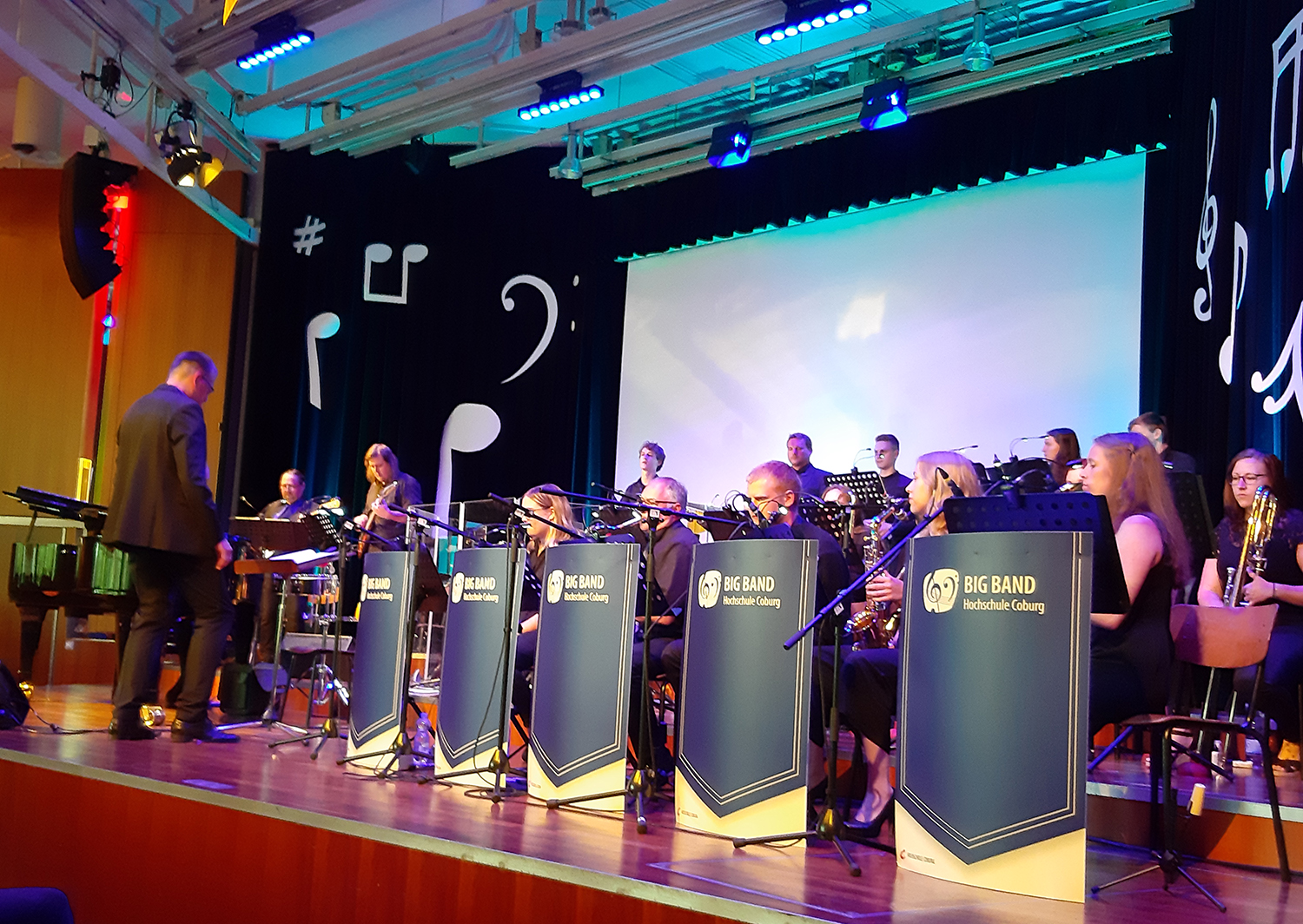 Bei einem Big-Band-Auftritt auf der Bühne spielen talentierte Musiker der Hochschule Coburg Saxophone und Trompeten. Sie sitzen hinter blauen Notenständern mit der Aufschrift „Big Band Hochschule Coburg“, umgeben von dekorativen Musiksymbolen.