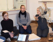 Eine Frau gestikuliert, während sie eine Skulptur mit baumelnden Buchstaben hält. Sie sitzt auf dem polierten Boden der Hochschule Coburg, während zwei Personen sie beobachten. Papiere und ein Laptop liegen verstreut um sie herum. Der Hintergrund besteht aus Holzboden und hellen Wänden, was eine Atmosphäre der Kreativität und des Lernens schafft.