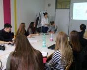 In einem modernen Raum mit bunt gestreiften Wänden sitzt eine Gruppe von Menschen um einen Konferenztisch. An der Hochschule Coburg steht eine Person ganz vorne und präsentiert Folien auf einer Leinwand. Die Teilnehmer machen sich Notizen und hören aufmerksam zu.
