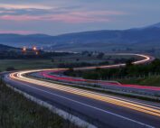 Eine Langzeitbelichtung einer Autobahn in der Abenddämmerung in der Nähe der Hochschule Coburg, bei der die Autolichter dynamische rote und weiße Streifen erzeugen. Die Straße schlängelt sich durch eine ländliche Landschaft mit sanften Hügeln im Hintergrund unter einem farbenfrohen Abendhimmel.
