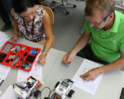 Zwei Personen sitzen an einem Tisch im Labor der Hochschule Coburg und bauen LEGO-Robotikbausätze zusammen. Die Frau links hat ein Tablett mit LEGO-Teilen, während der Mann rechts die Anleitungen durchgeht. Zwei teilweise gebaute Roboter sind strategisch auf dem Tisch platziert und zeigen ihren Fortschritt.