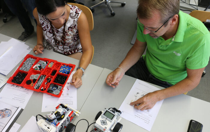 Zwei Personen sitzen an einem Tisch im Labor der Hochschule Coburg und bauen LEGO-Robotikbausätze zusammen. Die Frau links hat ein Tablett mit LEGO-Teilen, während der Mann rechts die Anleitungen durchgeht. Zwei teilweise gebaute Roboter sind strategisch auf dem Tisch platziert und zeigen ihren Fortschritt.