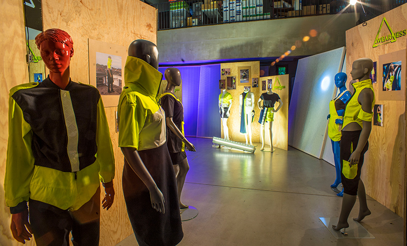 Im Ausstellungsraum der Hochschule Coburg sind Schaufensterpuppen in futuristischen, neonfarbenen Outfits zu sehen. Der Raum wird mit Scheinwerfern beleuchtet, die die leuchtenden Stoffe hervorheben, während die Wände mit entsprechenden Bildern und Displays geschmückt sind.