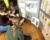 Drei Personen sitzen auf dem Holzboden eines Studios der Hochschule Coburg und diskutieren über Modeskizzen, die an der Wand hängen. Die Entwürfe zeigen Kleidung in Gelb- und Schwarztönen und schaffen eine kollaborative Atmosphäre, während sie gemeinsam kreative Ideen entwickeln.