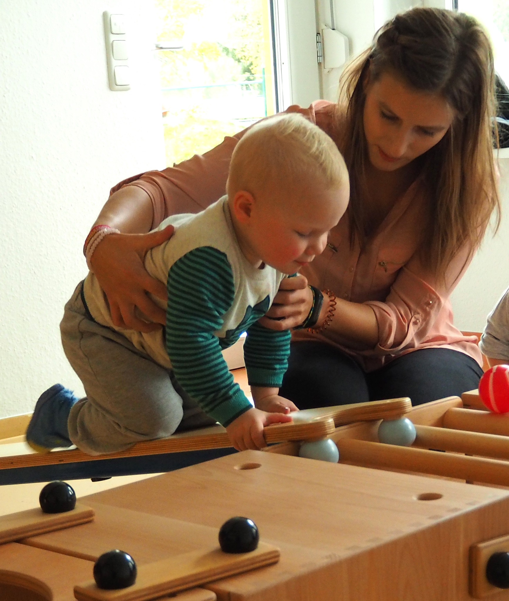 In einem sonnendurchfluteten Raum unterstützt eine Frau der Hochschule Coburg ein Baby beim Spielen mit einem Holzspielzeug mit Rampen und Bällen und sorgt so für wunderbare Momente im Haus.