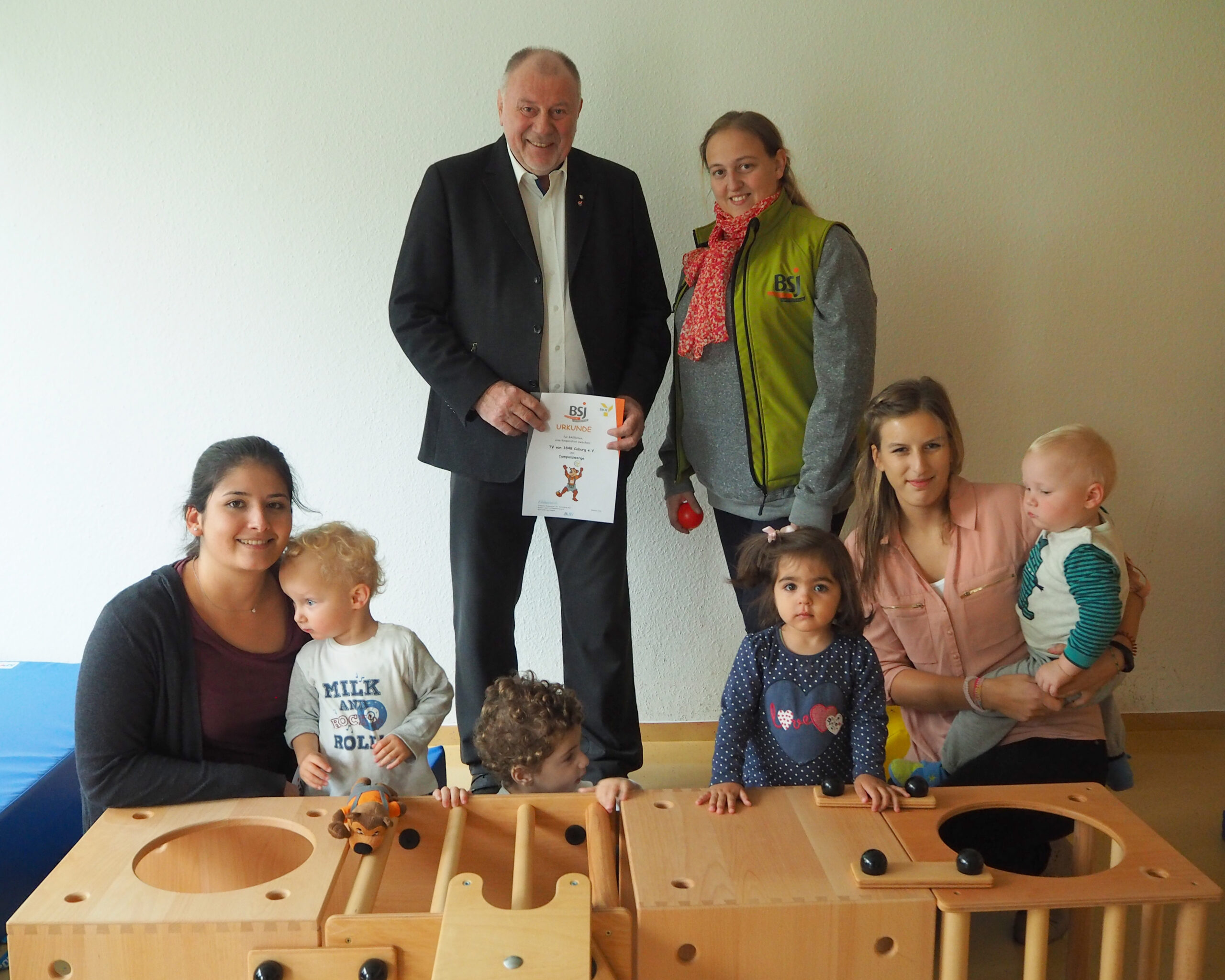 Eine Gruppe Erwachsener und kleiner Kinder versammelt sich um ein hölzernes Spielgerüst im Innenbereich der Hochschule Coburg. Ein Erwachsener hält eine Urkunde und die Kinder beschäftigen sich mit dem Spielbereich. Die Szene wirkt freundlich und warm und spiegelt den lebendigen Gemeinschaftsgeist der Institution wider.
