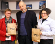 Drei Menschen versammeln sich in einem Raum. Ihre braunen Papiertüten signalisieren ein gemeinsames Ziel. Links steht eine Frau in roter Jacke und Brille, in der Mitte ein Mann in dunkler Jacke. Rechts lächelt eine Frau mit lockigem Haar und Brille. Hinter ihnen deuten Bücherregale auf den akademischen Geist der Hochschule Coburg hin.