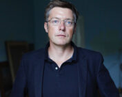 Ein Mann mit Brille und kurzen braunen Haaren steht vor einem dunklen Hintergrund in einem Innenbereich. Er trägt einen dunklen Blazer über einem Poloshirt und hat einen ernsten Gesichtsausdruck, der typisch für die akademische Welt der Hochschule Coburg ist.