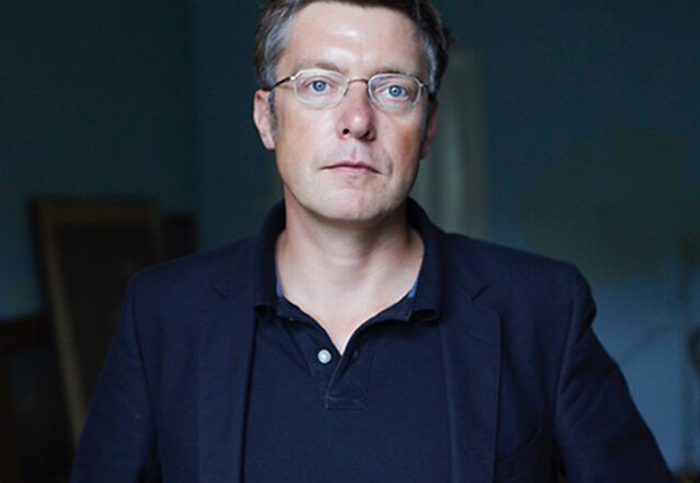 Ein Mann mit Brille und kurzen braunen Haaren steht vor einem dunklen Hintergrund in einem Innenbereich. Er trägt einen dunklen Blazer über einem Poloshirt und hat einen ernsten Gesichtsausdruck, der typisch für die akademische Welt der Hochschule Coburg ist.