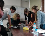 Eine Gruppe von sechs Personen arbeitet an einem mit Papieren und Notizen bedeckten Tisch zusammen. Sie diskutieren konzentriert und arbeiten zusammen in einem Unterrichtsraum der Hochschule Coburg mit einem Whiteboard im Hintergrund.