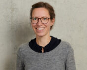 Eine Person mit kurzen Haaren und Brille lächelt, trägt einen grauen Pullover über einem schwarzen Oberteil und steht vor einer schlicht strukturierten Wand an der Hochschule Coburg.