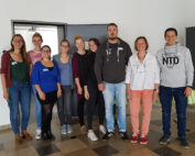 Eine Gruppe von neun Personen der Hochschule Coburg steht drinnen und lächelt in die Kamera. Sie sind leger gekleidet und stehen vor einer schlichten Wand. Einige halten kleine Taschen in den Händen. Der Boden unter ihnen ist mit einem gemischten Fliesenmuster versehen, was dem Ambiente einen Hauch von Charme verleiht.