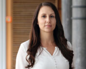 Eine Frau mit langen braunen Haaren steht im Innenbereich der Hochschule Coburg. Sie trägt eine weiße Bluse und blickt mit neutralem Gesichtsausdruck direkt in die Kamera. Der Hintergrund besteht aus Holz- und Metallelementen und fängt das moderne und doch warme Ambiente der Hochschule ein.