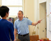 Ein Mann im hellblauen Poloshirt steht in einem Hörsaal der Hochschule Coburg. Er hält einen Zeigestab in der Hand und erklärt etwas auf einer Tafel voller Diagramme. Zwei Studierende sitzen ihm gegenüber und hören aufmerksam zu.