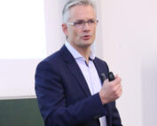 Ein Mann in blauem Anzug und Brille hält einen Vortrag an der Hochschule Coburg. Er hält eine Fernbedienung in der Hand und scheint vor einer Projektionsfläche zu sprechen.
