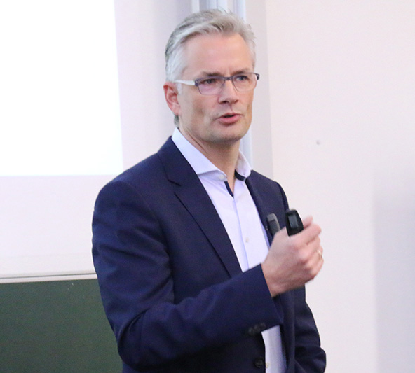 Ein Mann in blauem Anzug und Brille hält einen Vortrag an der Hochschule Coburg. Er hält eine Fernbedienung in der Hand und scheint vor einer Projektionsfläche zu sprechen.