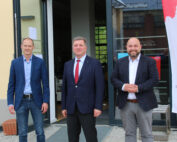 Drei Männer in Anzügen stehen lächelnd vor einem Gebäude mit einer Glastür. Der Mann links trägt Jeans und ein blaues Hemd, der Mann in der Mitte mit einer roten Krawatte hält Materialien der Hochschule Coburg in der Hand und der Mann rechts trägt ein weißes Hemd ohne Krawatte. Ein rot-weißes Schild ist teilweise sichtbar.