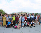 Eine große Gruppe von Studierenden und Lehrkräften der Hochschule Coburg posiert im Freien auf einer gepflasterten Fläche, im Hintergrund eine malerische Landschaft aus Bäumen und Hügeln. Der Himmel ist teilweise bewölkt. Die bunt gemischte Gruppe scheint auf einem Schulausflug oder einer Exkursion zu sein.