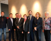 Eine Gruppe von acht Personen, die mit der Hochschule Coburg verbunden sind, steht Seite an Seite im Innenbereich. Sie tragen formelle und halbformelle Kleidung und lächeln herzlich vor einer schlichten Wand mit sanfter Beleuchtung darüber.