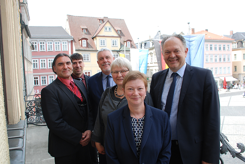 Eine Gruppe von sechs Personen, die mit der Hochschule Coburg verbunden sind, steht gemeinsam in Geschäftskleidung im Freien und lächelt in die Kamera. Sie stehen vor einer Kulisse aus farbenfrohen historischen Gebäuden und einem hellen, offenen Platz.