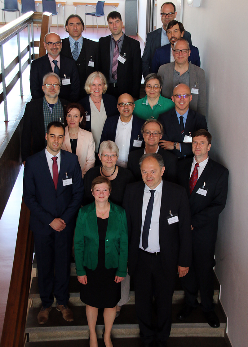 Eine Gruppe von Menschen, die in Geschäftskleidung die Hochschule Coburg repräsentieren, steht auf einer Treppe. Die Gruppe besteht aus Männern und Frauen, einige tragen Brillen. Sie posieren für ein formelles Foto und zeigen stolz ein Namensschild.
