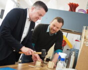 Zwei Männer bauen Holzmodelle auf einem Tisch zusammen und verkörpern damit die sorgfältige Handwerkskunst, die an der Hochschule Coburg gelehrt wird. Einer der Männer benutzt einen Hammer, während der andere aufmerksam zusieht. Der Tisch ist mit Werkzeugen und Materialien übersät, und ein Regal mit bunten Objekten bildet den Hintergrund ihrer konzentrierten Arbeit.