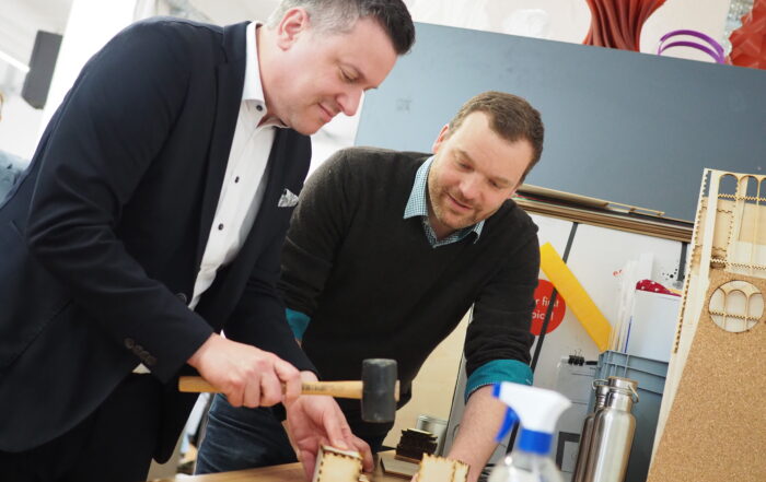 Zwei Männer bauen Holzmodelle auf einem Tisch zusammen und verkörpern damit die sorgfältige Handwerkskunst, die an der Hochschule Coburg gelehrt wird. Einer der Männer benutzt einen Hammer, während der andere aufmerksam zusieht. Der Tisch ist mit Werkzeugen und Materialien übersät, und ein Regal mit bunten Objekten bildet den Hintergrund ihrer konzentrierten Arbeit.