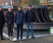 Vier Personen stehen auf einer gepflasterten Fläche vor einer großen, industriellen Skulptur, die einer Turbine ähnelt. Die Außenkulisse in der Nähe der Hochschule Coburg wird durch die umliegenden Gebäude, Bäume und Gras im Hintergrund aufgewertet.