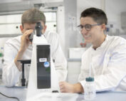 Zwei Menschen in Laborkitteln arbeiten gemeinsam im Labor der Hochschule Coburg. Einer blickt durch ein Mikroskop, der andere trägt eine Brille, lächelt und beobachtet. Auf dem Schreibtisch vor ihnen stehen eine Flasche und verschiedene Laborutensilien.