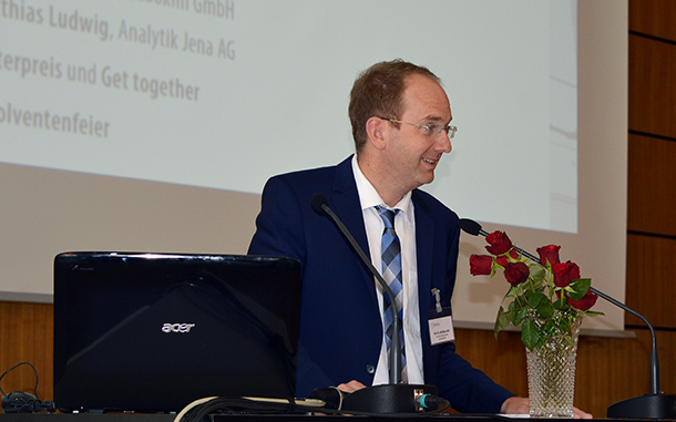 Eine Person im Anzug spricht während einer Veranstaltung an der Hochschule Coburg an einem Podium mit Mikrofon. Vor ihr steht ein aufgeklappter Laptop, an dessen Seite eine Vase mit roten Rosen steht. Im Hintergrund wird auf einer Leinwand eine Präsentation projiziert.
