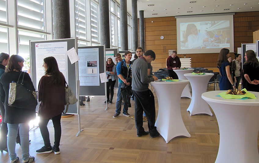 In einem Konferenzraum sind Menschen versammelt, die Präsentationsplakate ausstellen. An der Hochschule Coburg stehen mehrere Teilnehmer an Stehtischen, diskutieren und betrachten die Plakate. Im Hintergrund ist eine Projektionsleinwand zu sehen.