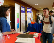 Eine Person steht an einem Stand und unterhält sich mit einem Teilnehmer der Veranstaltung. Der Stand der Hochschule Coburg ist mit farbenfrohen Bannern und Broschüren ausgestattet. Andere Personen stehen in der Nähe und schaffen eine lebendige Atmosphäre, die typisch für eine dynamische Konferenz- oder Ausstellungsumgebung ist.