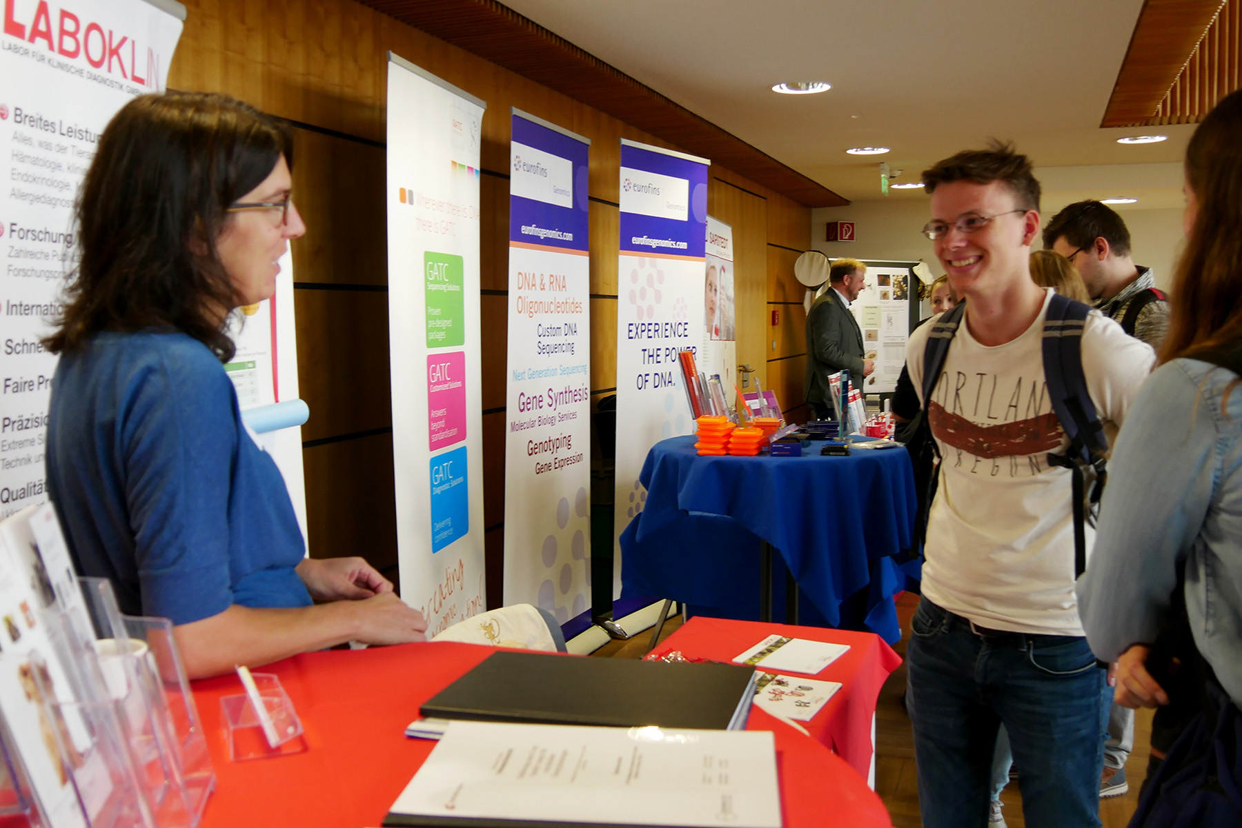 Eine Person steht an einem Stand und unterhält sich mit einem Teilnehmer der Veranstaltung. Der Stand der Hochschule Coburg ist mit farbenfrohen Bannern und Broschüren ausgestattet. Andere Personen stehen in der Nähe und schaffen eine lebendige Atmosphäre, die typisch für eine dynamische Konferenz- oder Ausstellungsumgebung ist.
