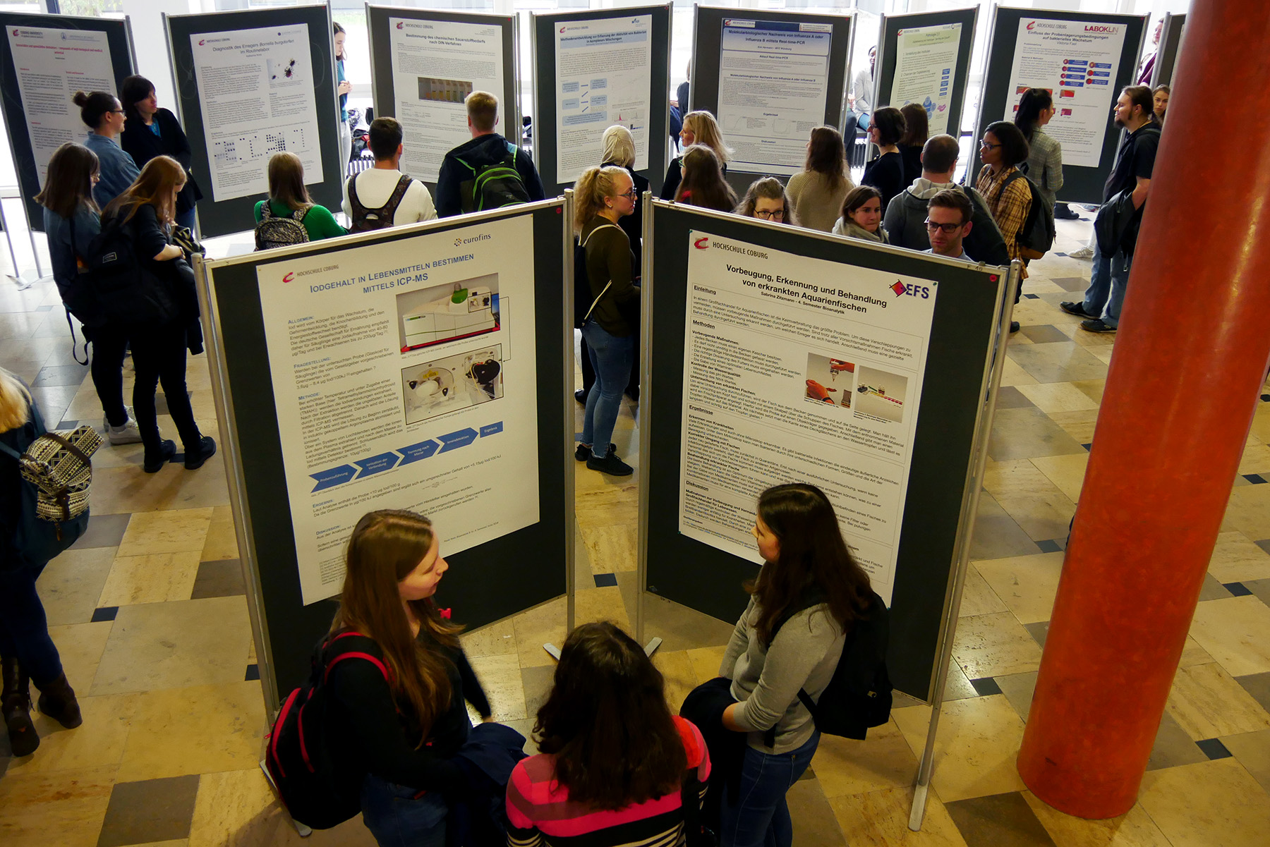 Eine Gruppe von Menschen besucht eine Postersession auf der Konferenz der Hochschule Coburg. Auf Tafeln werden zahlreiche wissenschaftliche Poster präsentiert, deren Inhalte die Teilnehmer lesen und diskutieren. Die Veranstaltung findet in einem geräumigen Innenbereich mit gefliestem Boden statt.