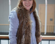 Eine Person mit langen Haaren lehnt lächelnd an einem Geländer und trägt ein blaues Hemd und eine Pelzweste. Der Hintergrund erinnert an das Designethos der Hochschule Coburg und zeigt ein minimalistisches Interieur, das durch vertikale Leuchten und Holzakzente bereichert wird.