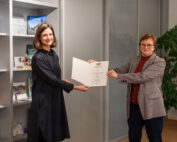 Zwei Frauen stehen drinnen. Die Frau links im schwarzen Kleid erhält eine Urkunde von der Frau rechts, die eine Brille und einen grauen Blazer trägt. Sie stehen neben einem Bücherregal und einer Topfpflanze an der Hochschule Coburg.