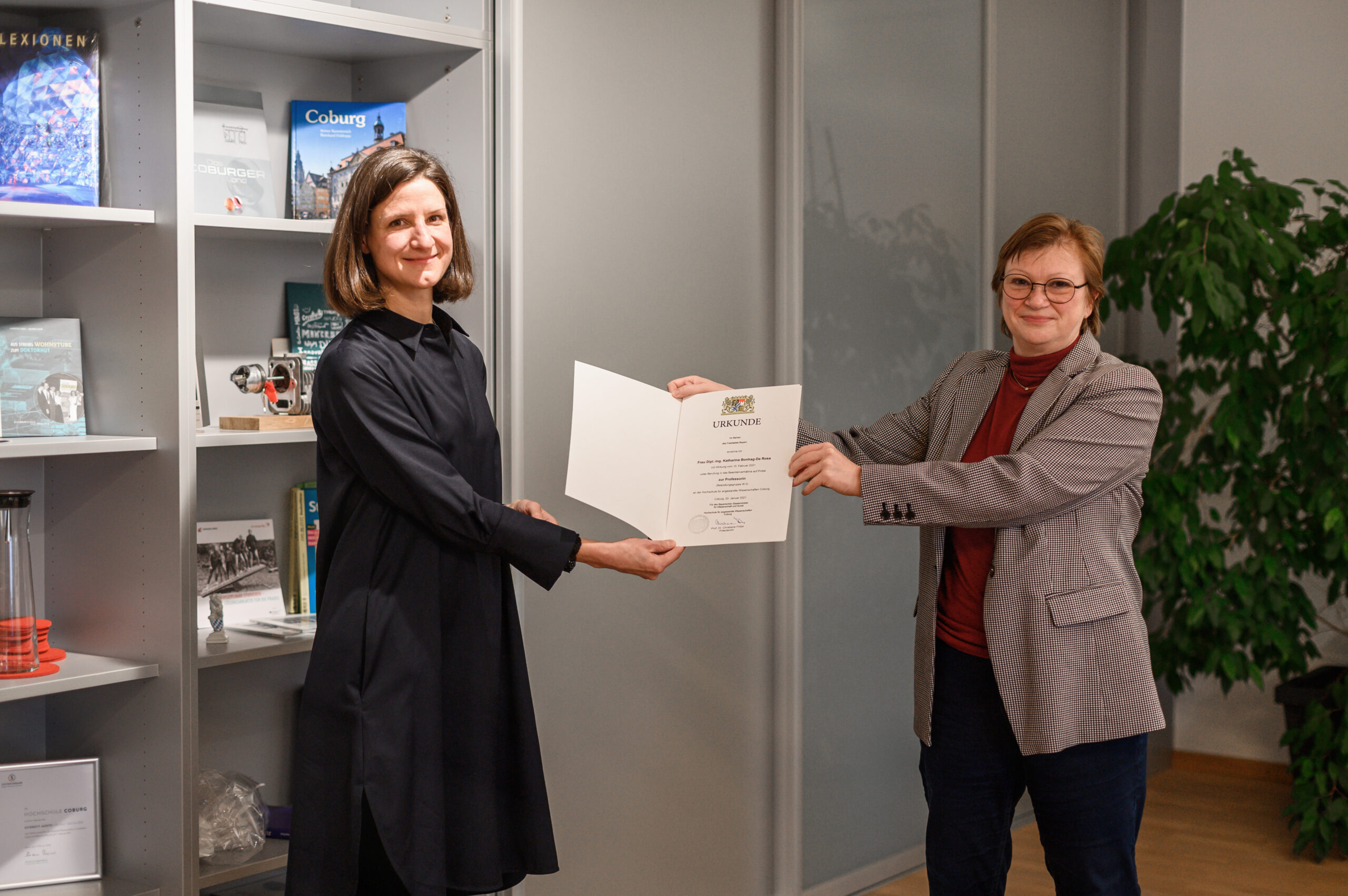 Zwei Frauen stehen drinnen. Die Frau links im schwarzen Kleid erhält eine Urkunde von der Frau rechts, die eine Brille und einen grauen Blazer trägt. Sie stehen neben einem Bücherregal und einer Topfpflanze an der Hochschule Coburg.