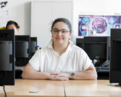 Eine Person mit Brille und weißem Hemd sitzt an einem Schreibtisch mit Computern in einem Büro oder einem Unterrichtsraum der Hochschule Coburg. Im Hintergrund ist eine weitere Person teilweise zu sehen. Ein lila Poster bringt Farbe an die Wand.