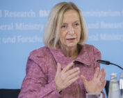 Eine Person mit blondem Haar spricht auf einer Pressekonferenz der Hochschule Coburg und gestikuliert lebhaft in einer rosa Jacke. Der verschwommene Text auf der blauen Wand weist auf das Thema der Veranstaltung hin. Zwei Mikrofone und eine Wasserflasche stehen prominent auf dem Tisch, während sie das Publikum anspricht.