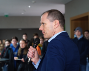 Ein Mann im blauen Anzug hält ein Mikrofon in der Hand und spricht zu einer Gruppe an der Hochschule Coburg. Die leger gekleideten Teilnehmer hören aufmerksam zu, was darauf schließen lässt, dass es sich hier um ein Fachseminar oder eine Bildungsveranstaltung handelt.