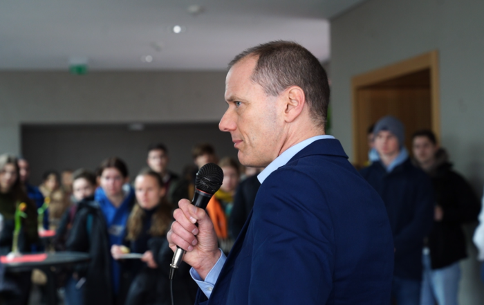 Ein Mann im blauen Anzug hält ein Mikrofon in der Hand und spricht zu einer Gruppe an der Hochschule Coburg. Die leger gekleideten Teilnehmer hören aufmerksam zu, was darauf schließen lässt, dass es sich hier um ein Fachseminar oder eine Bildungsveranstaltung handelt.